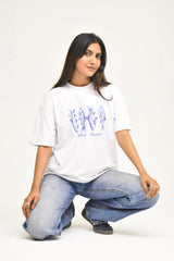 A woman crouching in a studio setting, wearing a white oversized graphic T-shirt with blue fish illustrations and the text 'Khaar Studios' underneath, paired with light-wash blue jeans.