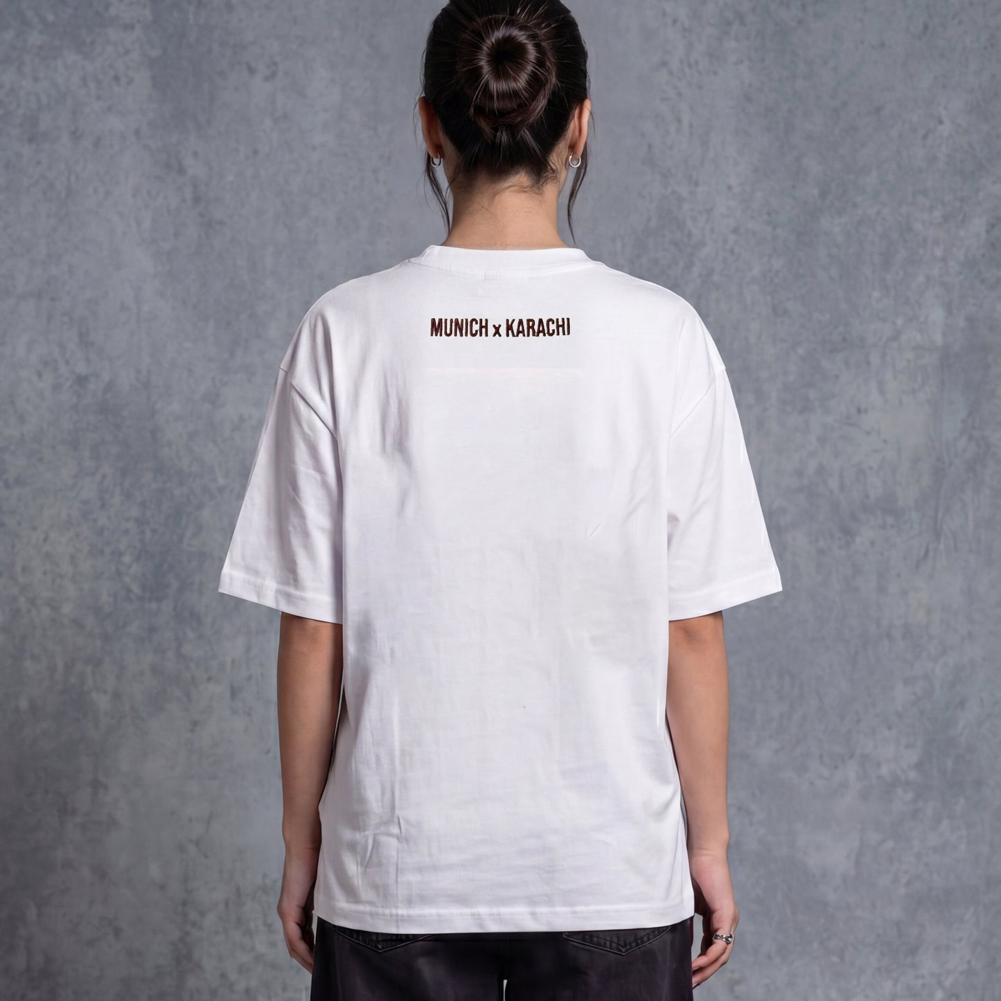 A person shown from the back wearing a white oversized T-shirt with blue 'MINCH + KARACHI' text on the upper back, paired with black shorts, white socks, and dark sneakers, standing against a textured gray studio backdrop.